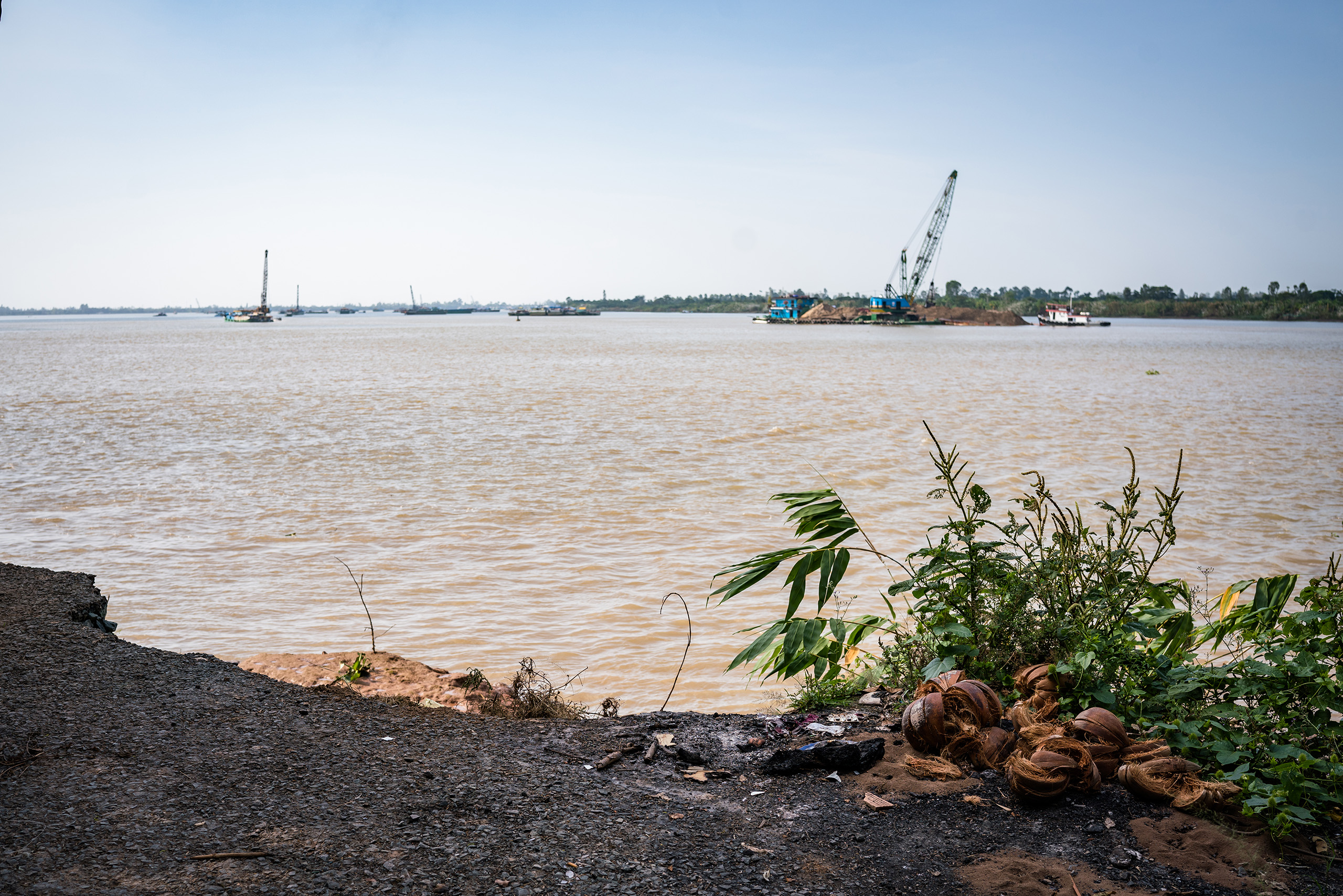 A road on Tan Thuan Dong island ends abruptly where it has been washed away. In the background, excavators dredge the river for sand
