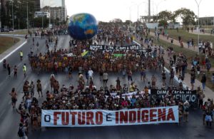 <p>Indigenous people protest against Brazil’s president, Jair Bolsonaro, and in favour of the demarcation of indigenous lands, in Brasília, April 2022. The group carries a banner with the words ‘The future is indigenous’. (Image: Adriano Machado / Alamy)</p>