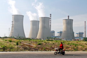 <p>A woman rides her electric bike past a coal-fired power plant in Huaian city (Image: David Lyons / Alamy)</p>
