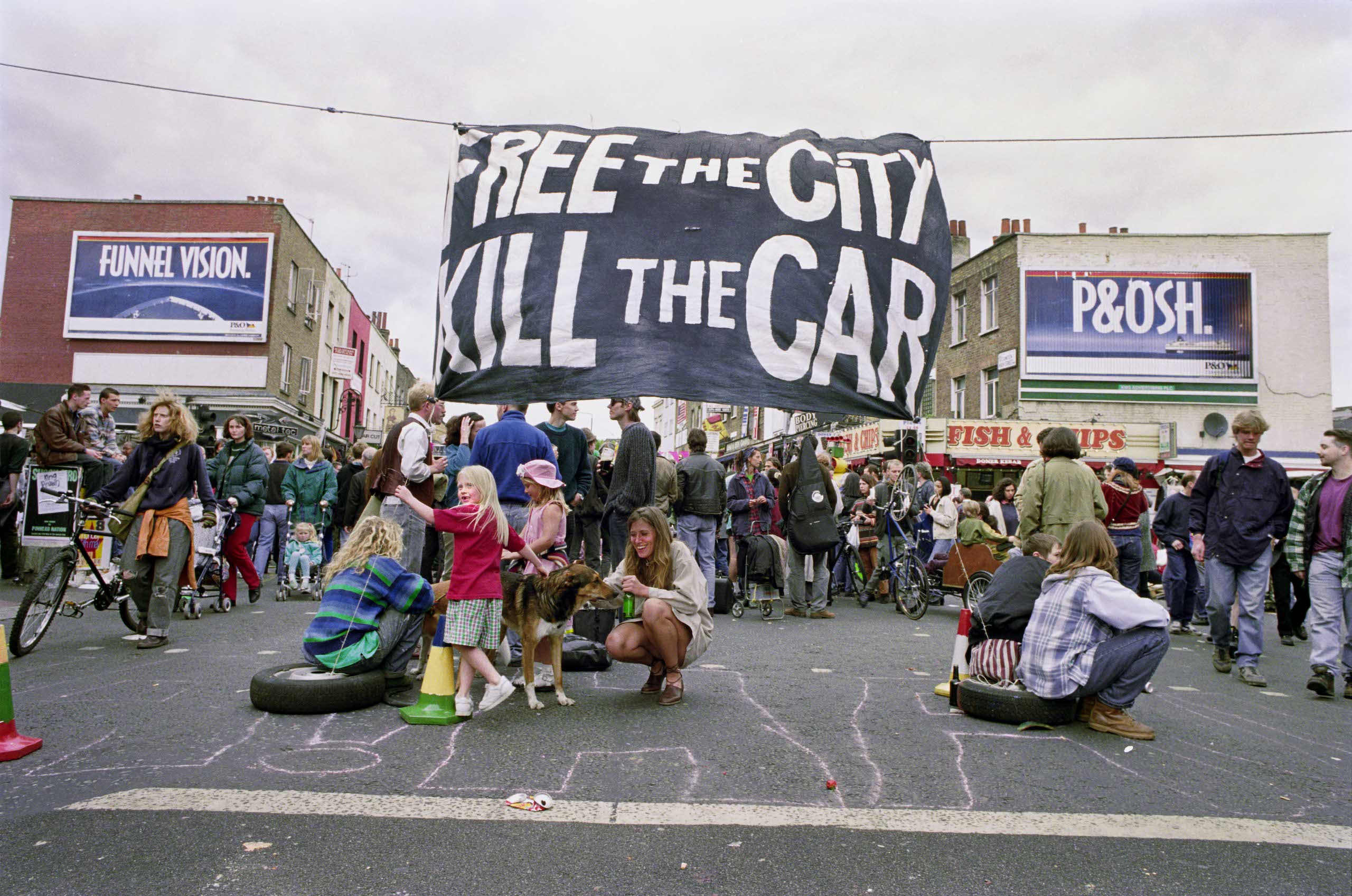 A large black and white banner hangs between buildings: Free the City, Kill the car. People sit in the road protesting and talking