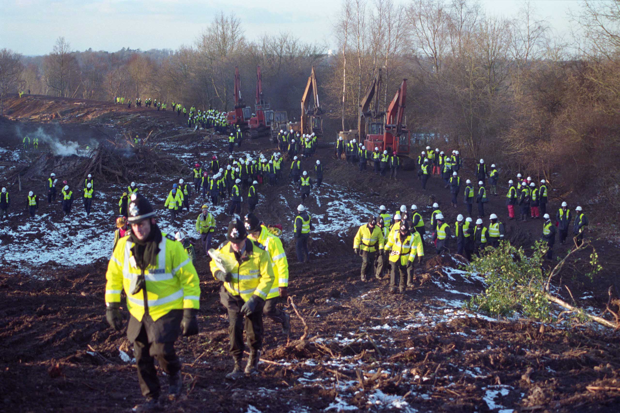 A long line of police in high vis jackets walk over mud and by trees