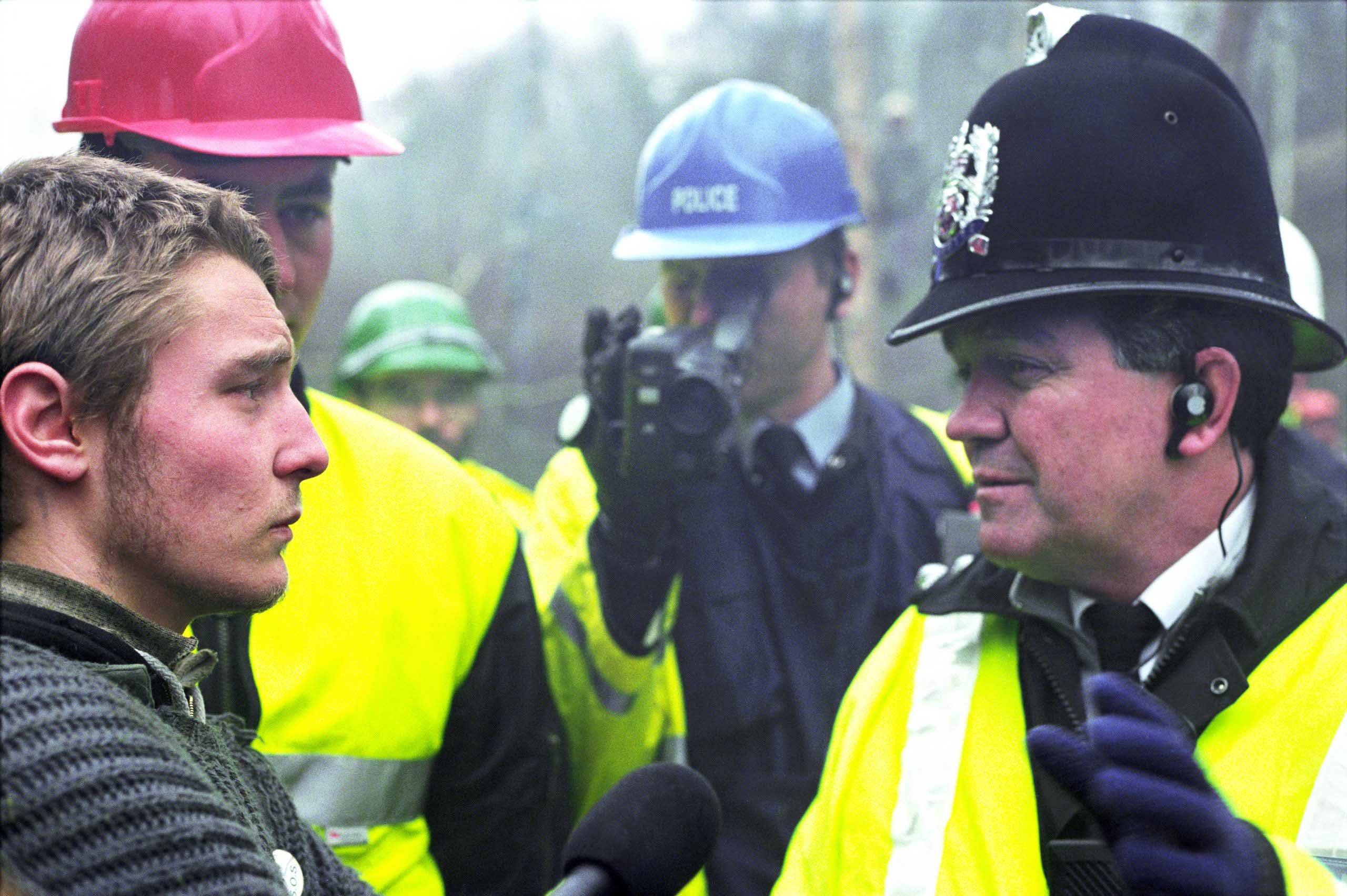 A young man makes eye contact with a police officer while someone films on a video camera in the background. Historical photograph