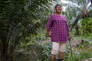 A woman dressed in pink holding a spray nozzle, stands next to a bushy green oil palm tree