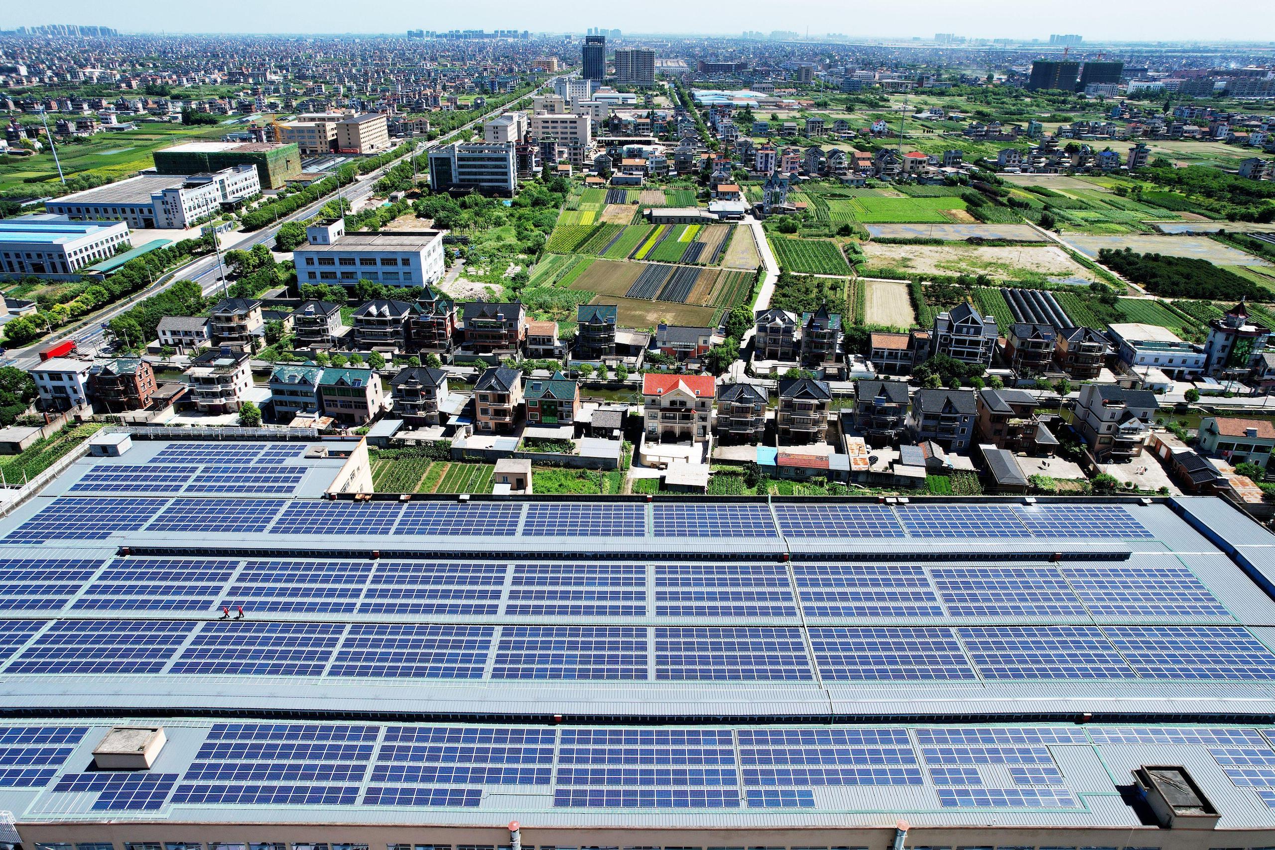 Hundreds of solar panels on the roof of a factory, aerial photo, Chinese small town countryside view in background, green rice fields and mid-rise buildings