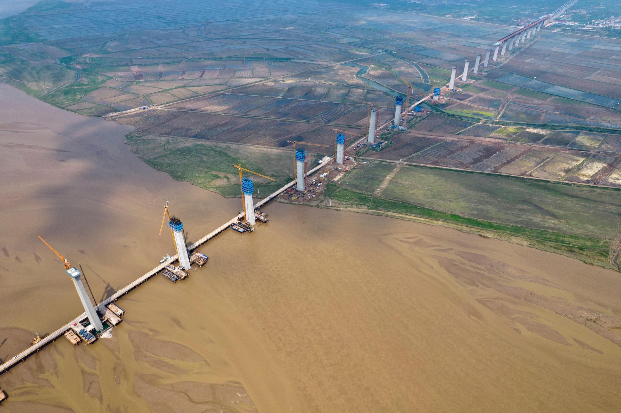 Aerial photo shows the construction site of Linyi Yellow River Bridge, strip of grey concrete pillars in a line across rural landscape, yellow silty water of the river, green agricultural fields