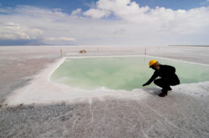Man wearing yellow hard hat dips hand in turquoise brine pool, backdrop of Salar de Uyuni salt flats in Bolivia, flat white desert scenery