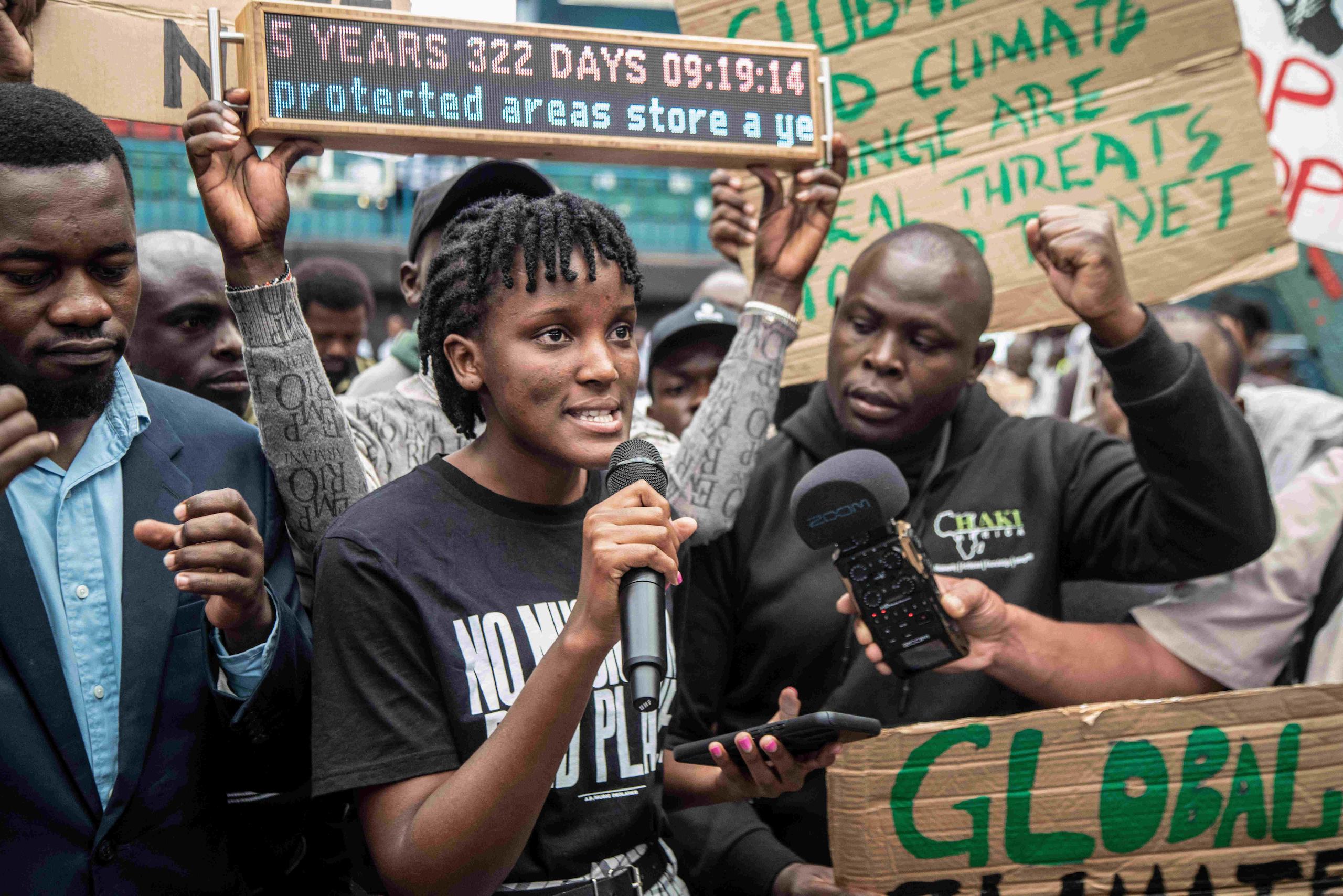 Woman speaks into a microphone surrounded by a crowd of people holding signs and raising their hands